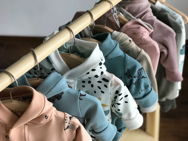 Różnokolorowe bluzy dziecięce z kapturami, wiszące na drewnianym stojaku z metalowymi wieszakami, prezentujące wzory zwierzęce i abstrakcyjne.