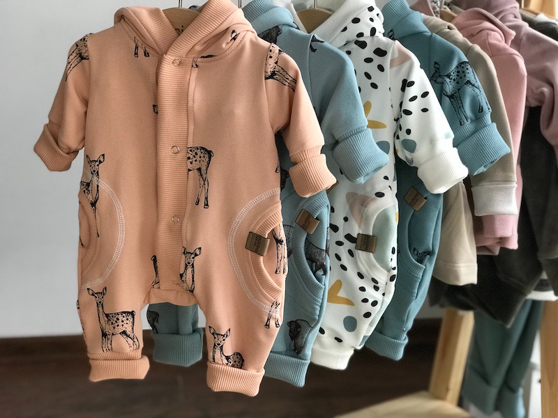 Rząd kolorowych kombinezonów dziecięcych z kapturem, z nadrukiem jelonków i geometrycznych wzorów, wiszących na drewnianym wieszaku.