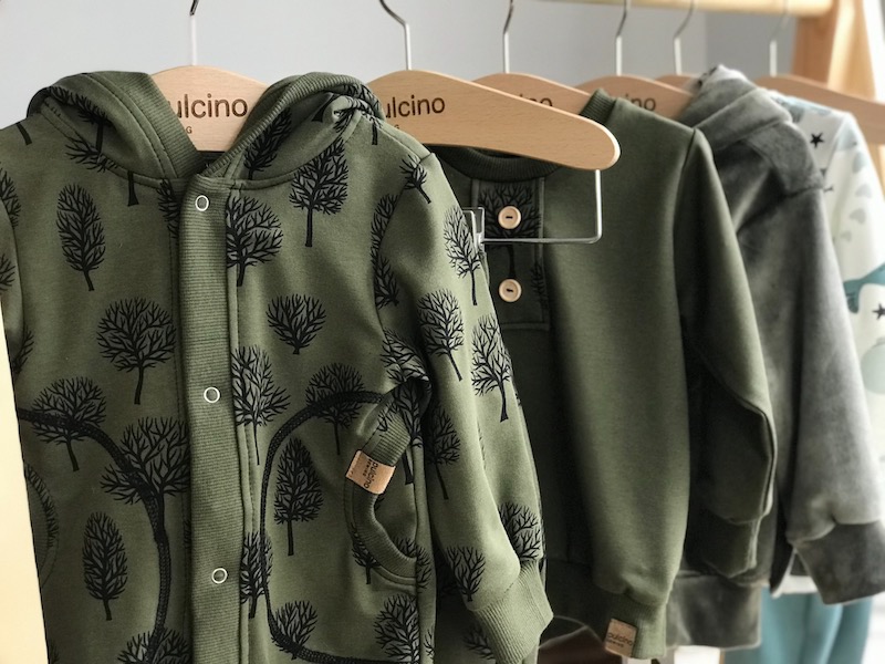 Rząd ubrań dziecięcych w odcieniach zieleni i szarości, wiszących na drewnianym wieszaku, w tym bluza z kapturem w drzewka i sweter zapinany na guziki.