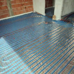 Niebieskie rury ogrzewania podłogowego ułożone na folii z czerwoną siatką w surowym wnętrzu z ceglanymi i betonowymi ścianami.