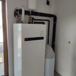 Biała pompa ciepła Panasonic z osprzętem (rury, zbiornik wyrównawczy, zawory) w surowym pomieszczeniu, widoczne czarne rury w izolacji i zmiękczacz wody obok pompy.