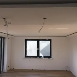 Wnętrze w trakcie remontu: świeżo położona jasna podłoga, białe ściany z widocznymi śladami po pracach, wystające kable z sufitu, okno z zabrudzeniami budowlanymi, widoczny fragment osoby po prawej...