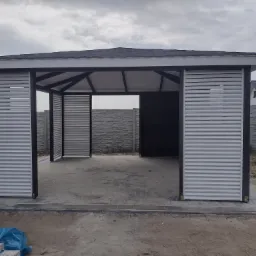 Nowoczesna, otwarta altana z szarymi, poziomymi panelami żaluzjowymi, czarną konstrukcją i dachówką bitumiczną, widoczna na tle szarego, betonowego ogrodzenia i pochmurnego nieba. Na pierwszym...