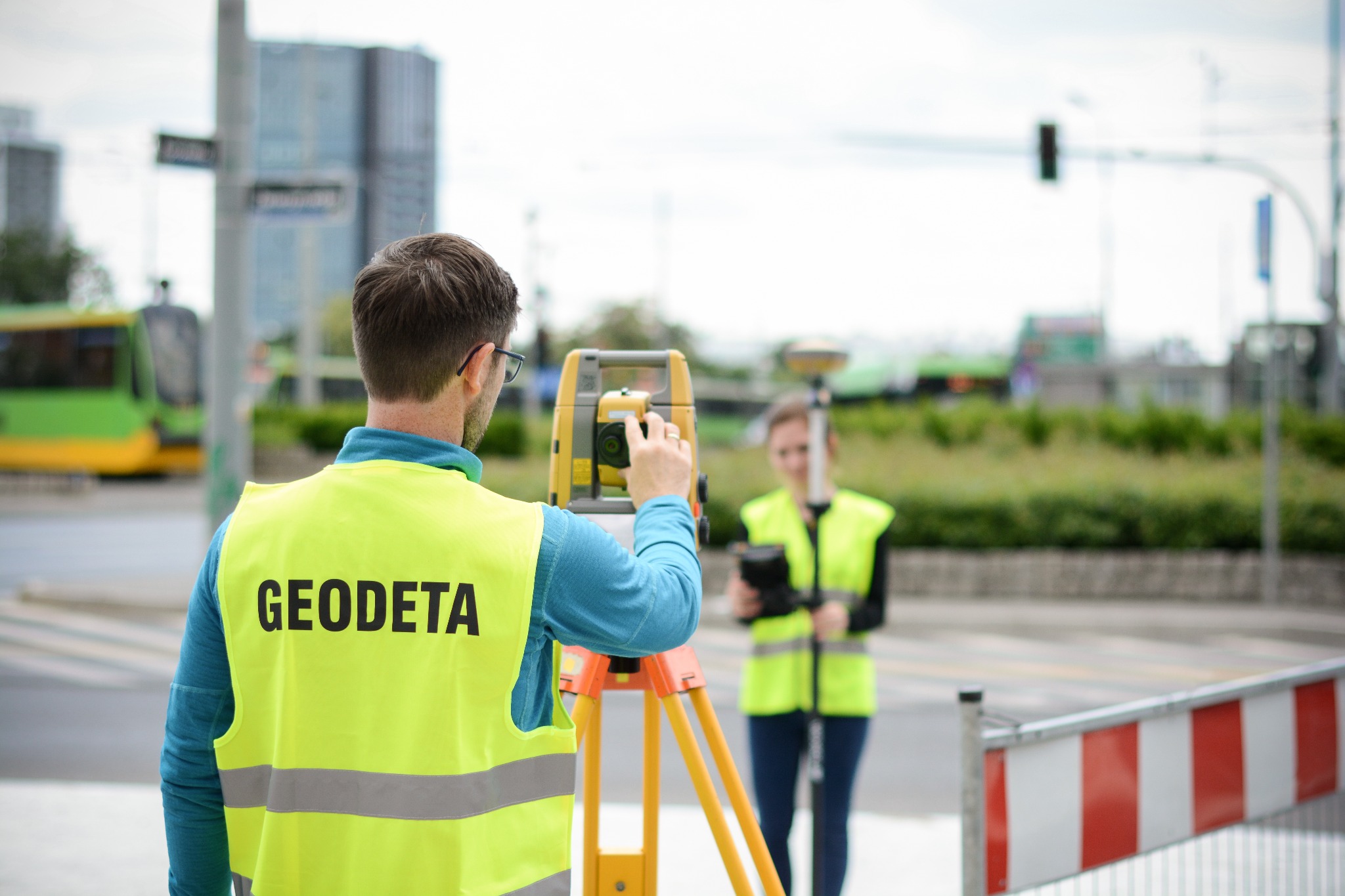 Dwóch pracowników w żółtych kamizelkach ochronnych, jeden celuje żółtym teodolitem, w tle miejski krajobraz z tramwajem i wieżowcami.