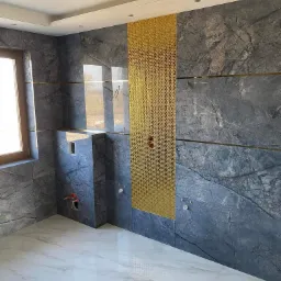 Wnętrze łazienki w trakcie wykańczania: ściany wyłożone szarym marmurem z złotymi listwami i dekoracyjną złotą mozaiką, biała marmurowa podłoga, widoczne instalacje wodne i okno.