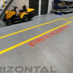 Horizontal Paweł Kasner - Oznakowana żółtymi liniami strefa rozładunku z czerwonym napisem na szarym betonowym podłożu w hali magazynowej, obok stoi kosiarka samojezdna.
