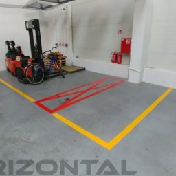 Horizontal Paweł Kasner - Oznakowanie poziome w magazynie: żółte linie wyznaczające strefy ruchu i czerwone X wewnątrz narożnika, w tle wózek widłowy i rower oparty o paletę.