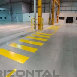 Horizontal Paweł Kasner - Żółte pasy namalowane na podłodze hali produkcyjnej w Rybniku, wyznaczające przejście dla pieszych i strefy bezpieczeństwa. Widoczne słupy konstrukcyjne i budynek w tle.