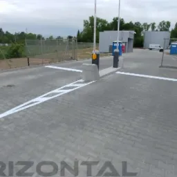 Oznakowana, betonowa droga dojazdowa z szlabanem, białe pasy i strzałki na szarym bruku, w tle budynki przemysłowe i zieleń.