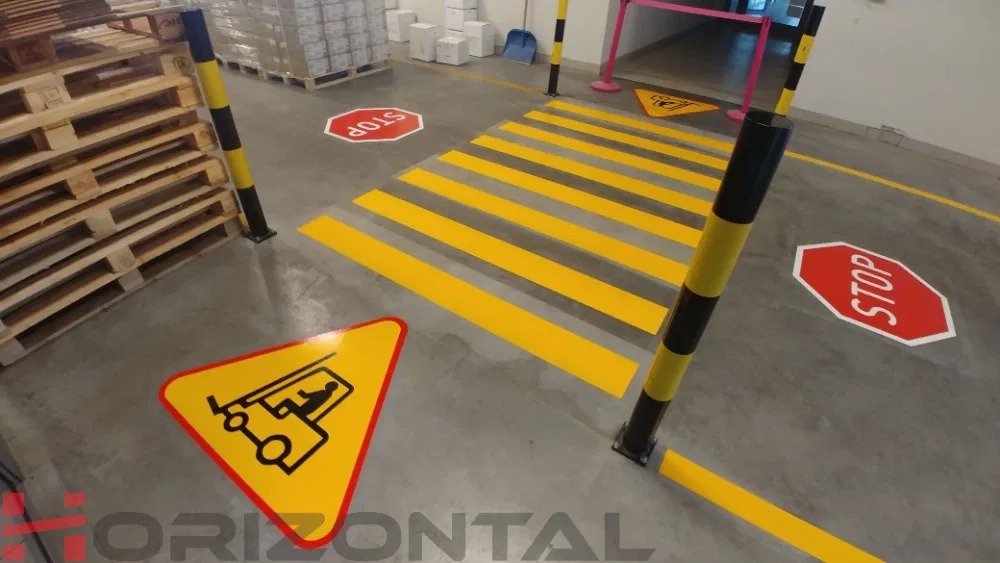 Oznakowanie poziome w hali magazynowej: żółte pasy przejścia dla pieszych, znaki ostrzegawcze z symbolem wózka widłowego, znaki STOP, słupki ochronne w czarno-żółte pasy.