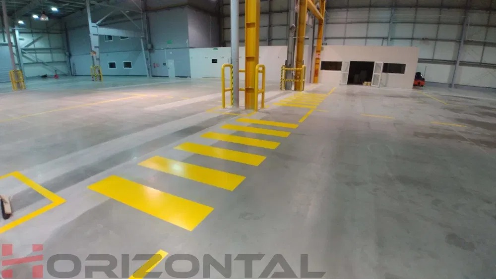 Żółte pasy namalowane na podłodze hali produkcyjnej w Rybniku, wyznaczające przejście dla pieszych i strefy bezpieczeństwa. Widoczne słupy konstrukcyjne i budynek w tle.