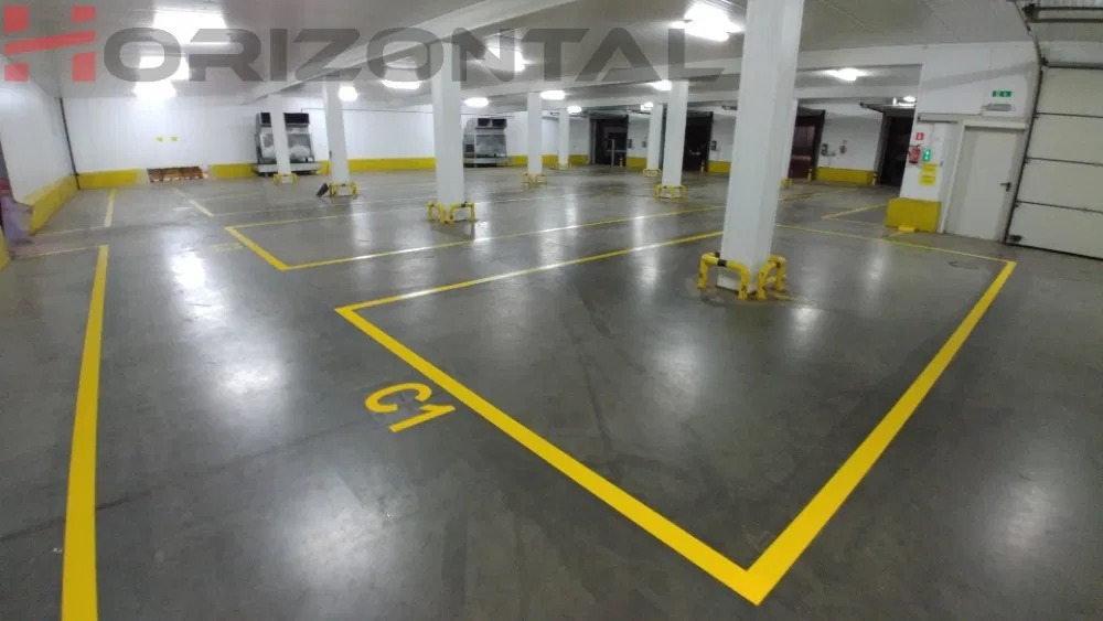 Oznakowane żółtą farbą stanowiska parkingowe w garażu podziemnym, z numerem 'C1' na jednym z nich, widoczne białe filary i żółte zabezpieczenia.