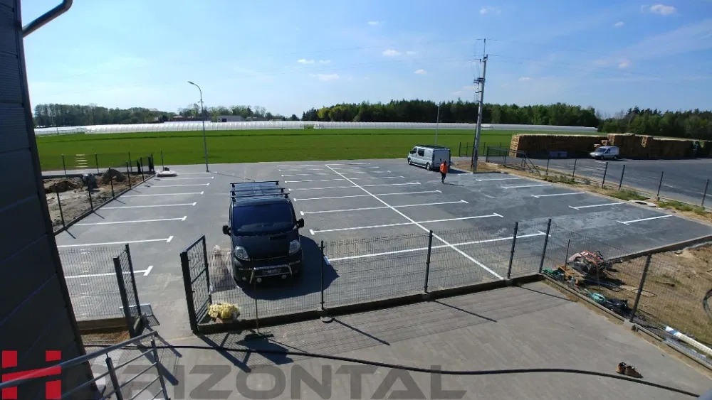 Widok z góry na nowo wybudowany parking z wyznaczonymi białymi liniami stanowisk, zaparkowany czarny bus z bagażnikiem dachowym, w tle zielone pole i hala.