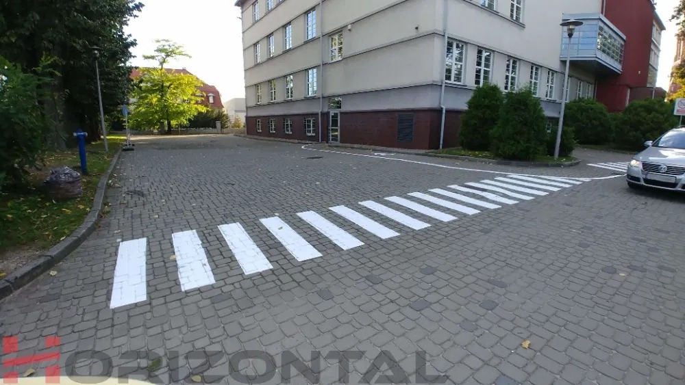 Nowo wymalowane białe pasy na przejściu dla pieszych, na kostce brukowej, przed budynkiem, z samochodem w tle.