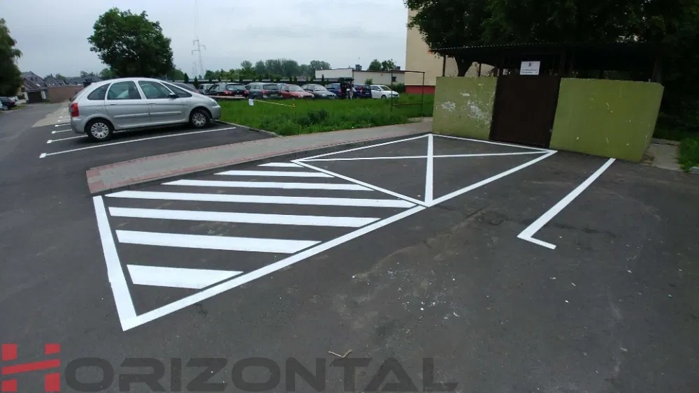 Oznakowanie poziome na parkingu: świeżo wymalowane pasy przejścia dla pieszych i miejsca parkingowe, widoczny srebrny samochód i budynek w tle.