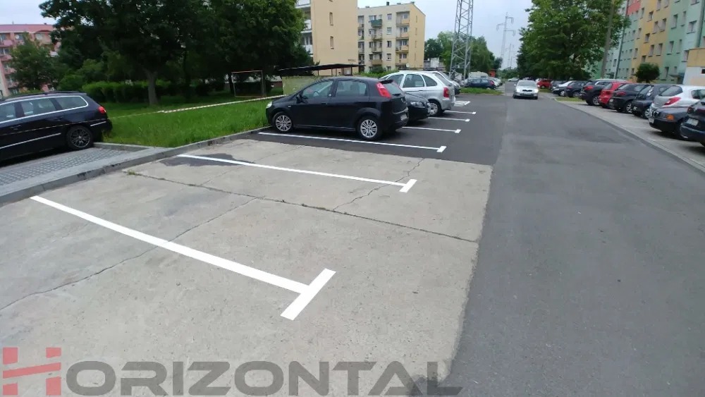 Oznakowane, białe linie na betonowym parkingu przed blokiem mieszkalnym, samochody zaparkowane wzdłuż linii, widok z perspektywy ulicy.