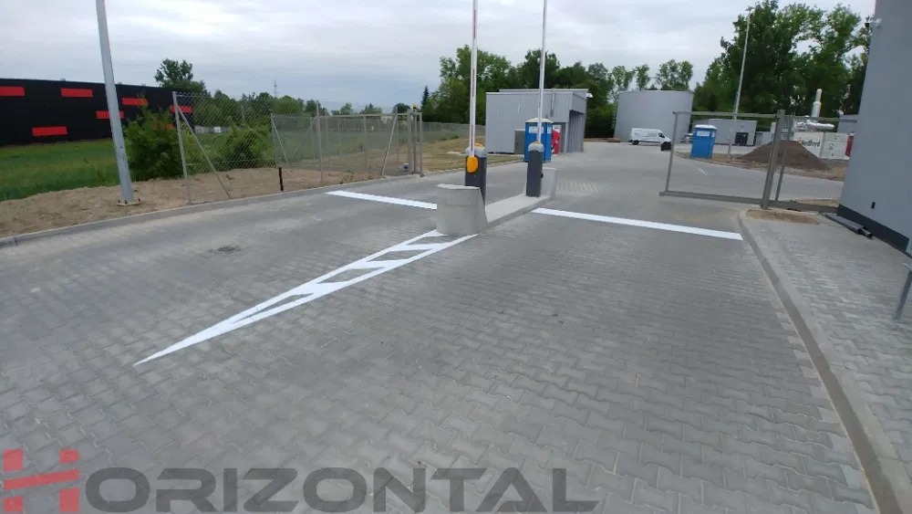 Oznakowana, betonowa droga dojazdowa z szlabanem, białe pasy i strzałki na szarym bruku, w tle budynki przemysłowe i zieleń.