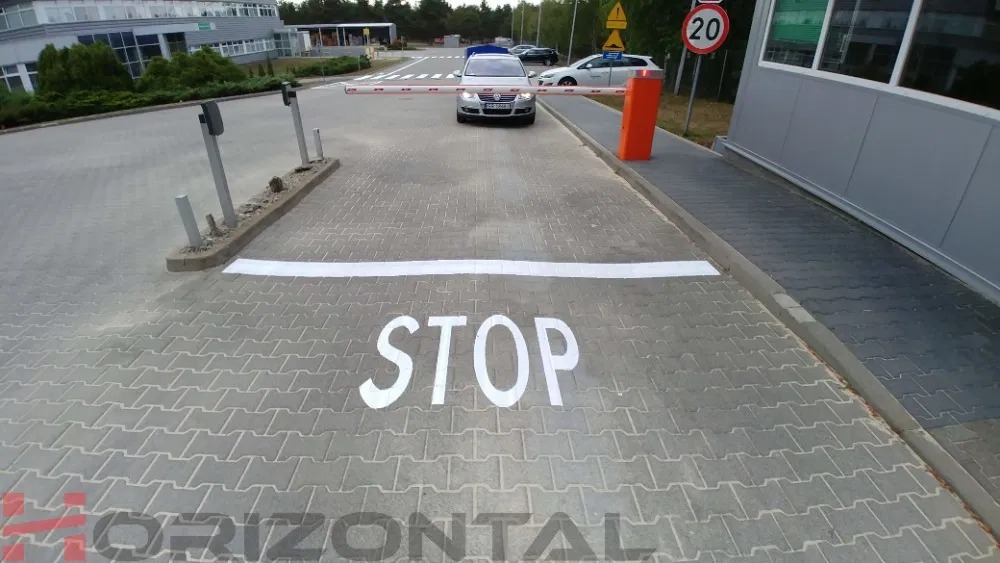 Biały napis 'STOP' i linia na szarej kostce brukowej przed szlabanem parkingowym, z samochodem w tle.