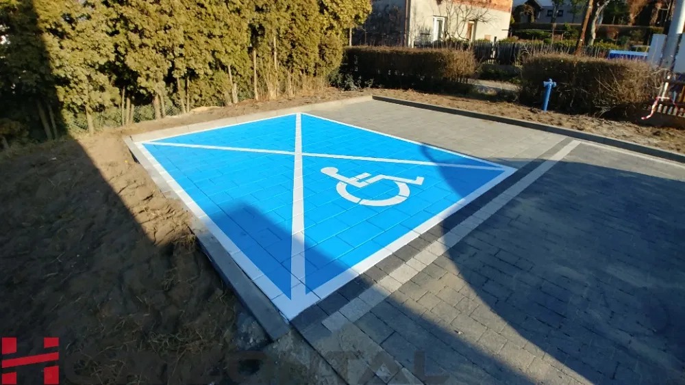 Oznakowane niebieskie miejsce parkingowe dla osób niepełnosprawnych, wykonane z kostki brukowej, otoczone szarą kostką i ziemią z roślinnością.