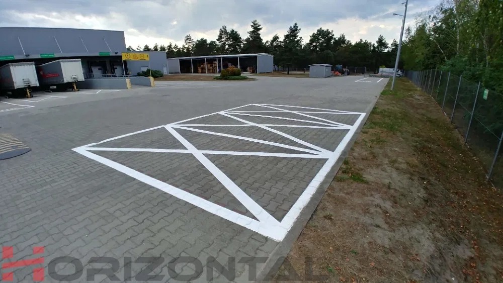 Nowo pomalowane, białe oznakowanie poziome na parkingu z kostki brukowej przed budynkiem magazynowym. Linie tworzą geometryczny wzór ograniczający miejsca parkingowe.