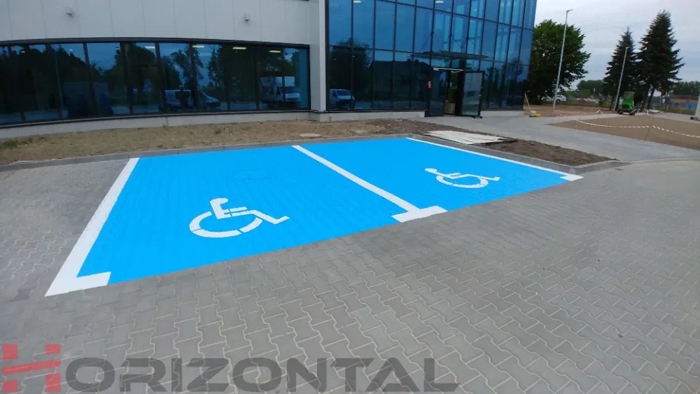Dwa nowo pomalowane, niebieskie miejsca parkingowe dla osób niepełnosprawnych z białymi symbolami i liniami, na szarej kostce brukowej, przed nowoczesnym budynkiem z przeszkloną fasadą.