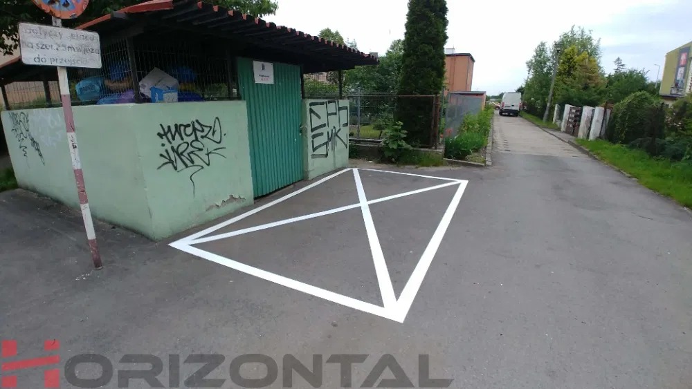 Oznakowane białą farbą miejsce parkingowe na asfalcie, obok śmietnika z graffiti, w tle droga i zieleń.