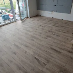 Układanie paneli podłogowych o wzorze drewna w jasnym pomieszczeniu z dużym oknem balkonowym i szarą ścianą częściowo wykończoną panelami dekoracyjnymi, widoczne narzędzia i materiały budowlane...