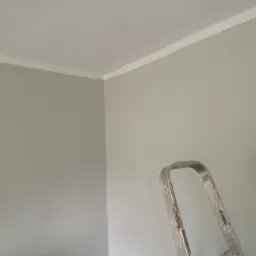 Świeżo pomalowane szare ściany z białą listwą pod sufitem, widoczny fragment drabiny z zabrudzeniami farby.
