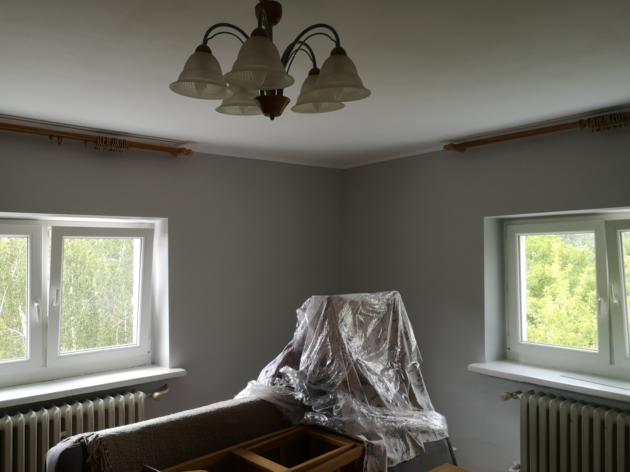 Pokój przed odświeżeniem: szare ściany, meble przykryte folią ochronną, widoczne grzejniki pod oknami, żyrandol na suficie.