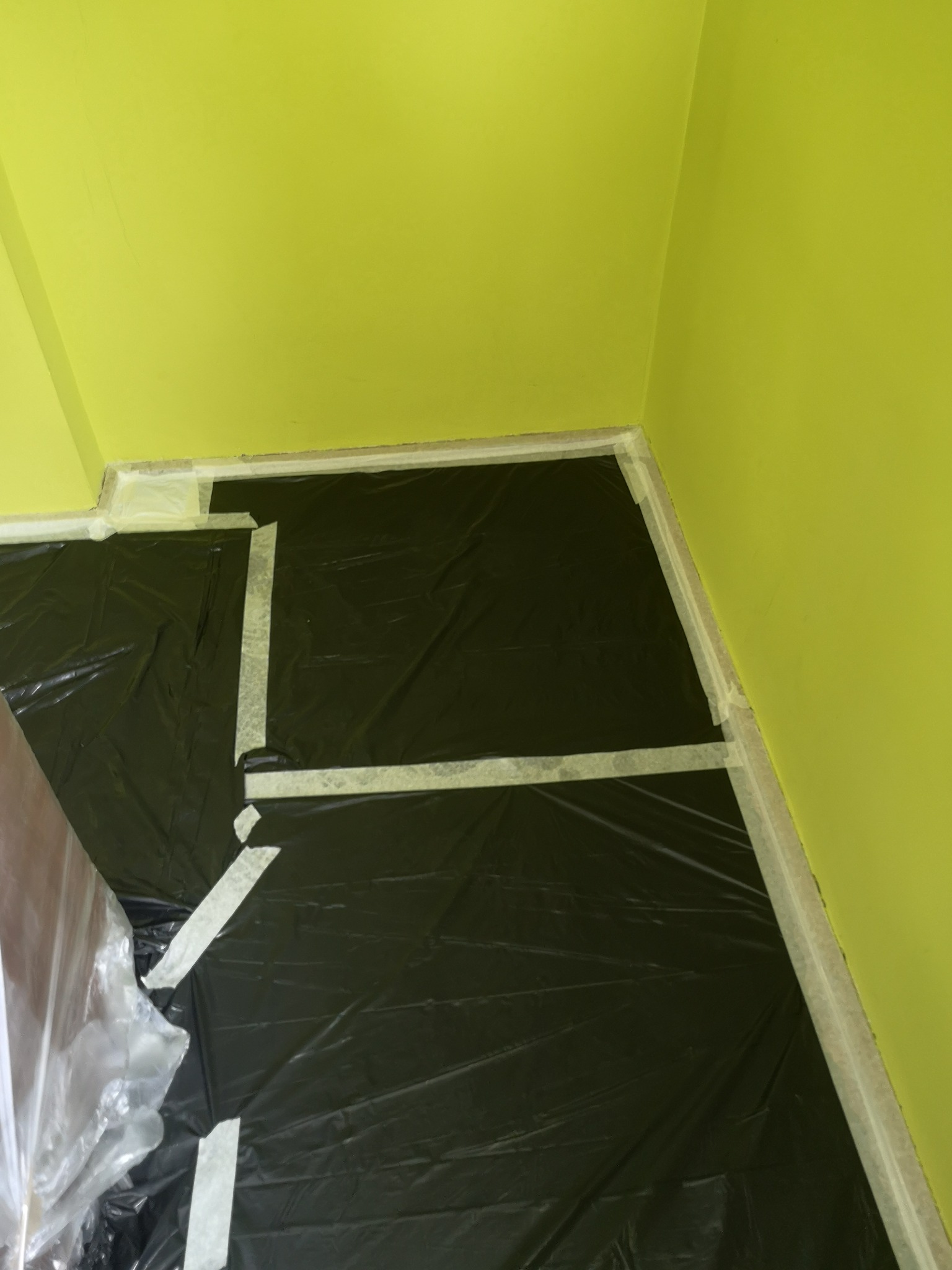 Przygotowanie do prac malarskich: podłoga zabezpieczona czarną folią i taśmą malarską przy ścianach pomalowanych na jasnozielono.