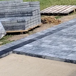 Układanie szarej kostki brukowej z czarnym obramowaniem, stos kostki na palecie w tle, piasek przygotowany pod dalsze prace.