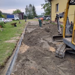 SM-Bud Brukarstwo - Pracownik wyrównuje ziemię łopatą wzdłuż krawężnika, w tle żółta ściana budynku i minikoparka, przygotowanie terenu pod budowę.