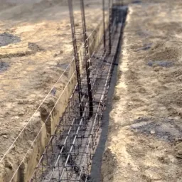 Zbrojenie ławy fundamentowej w wykopie, widoczne wystające pręty pionowe, w tle drzewa i budynek mieszkalny.