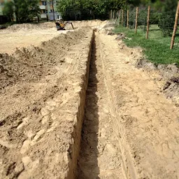 Wąski wykop liniowy w piaszczystej glebie, widoczny ślad po sznurze wyznaczającym linię, w tle mała koparka i budynek mieszkalny.