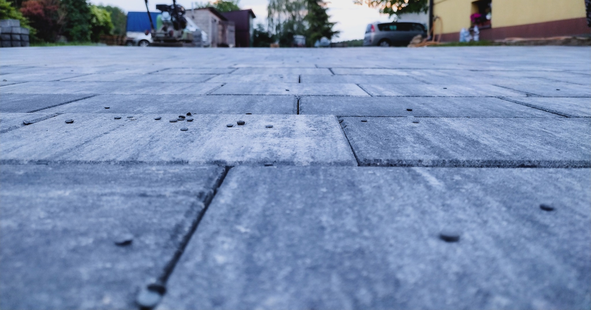 Niski kąt widzenia na szare kostki brukowe z widocznymi drobnymi elementami wypełniającymi szczeliny, w tle rozmyty krajobraz z budynkami i pojazdami.