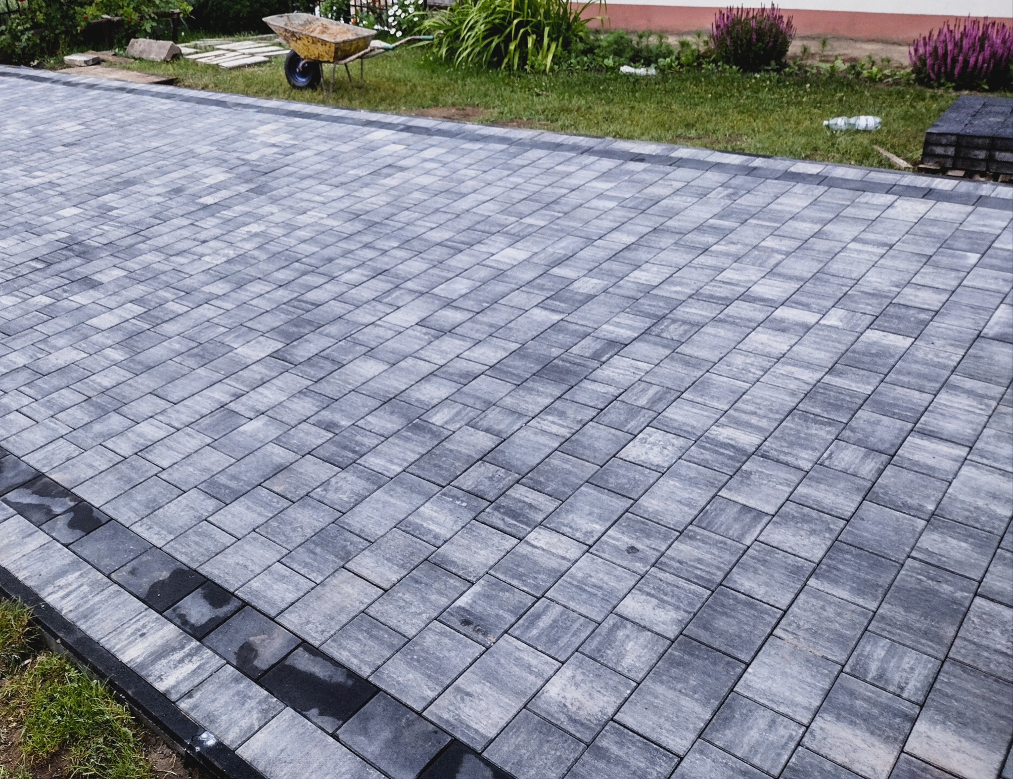 Świeżo ułożona, szara kostka brukowa z ciemnym obramowaniem na posesji we Władysławowie, z widocznym w tle wózkiem budowlanym i zielenią.