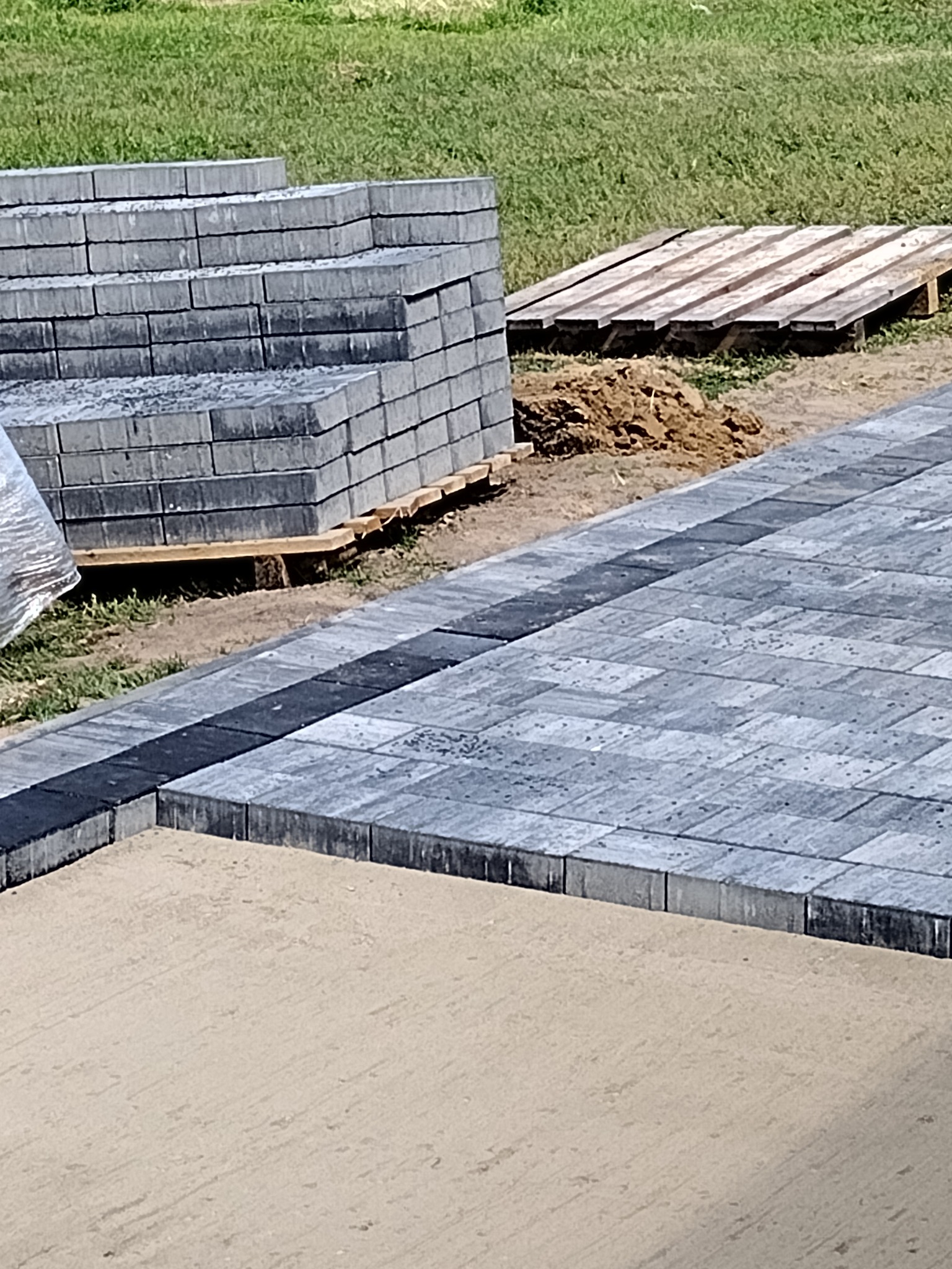 Układanie szarej kostki brukowej z czarnym obramowaniem, stos kostki na palecie w tle, piasek przygotowany pod dalsze prace.