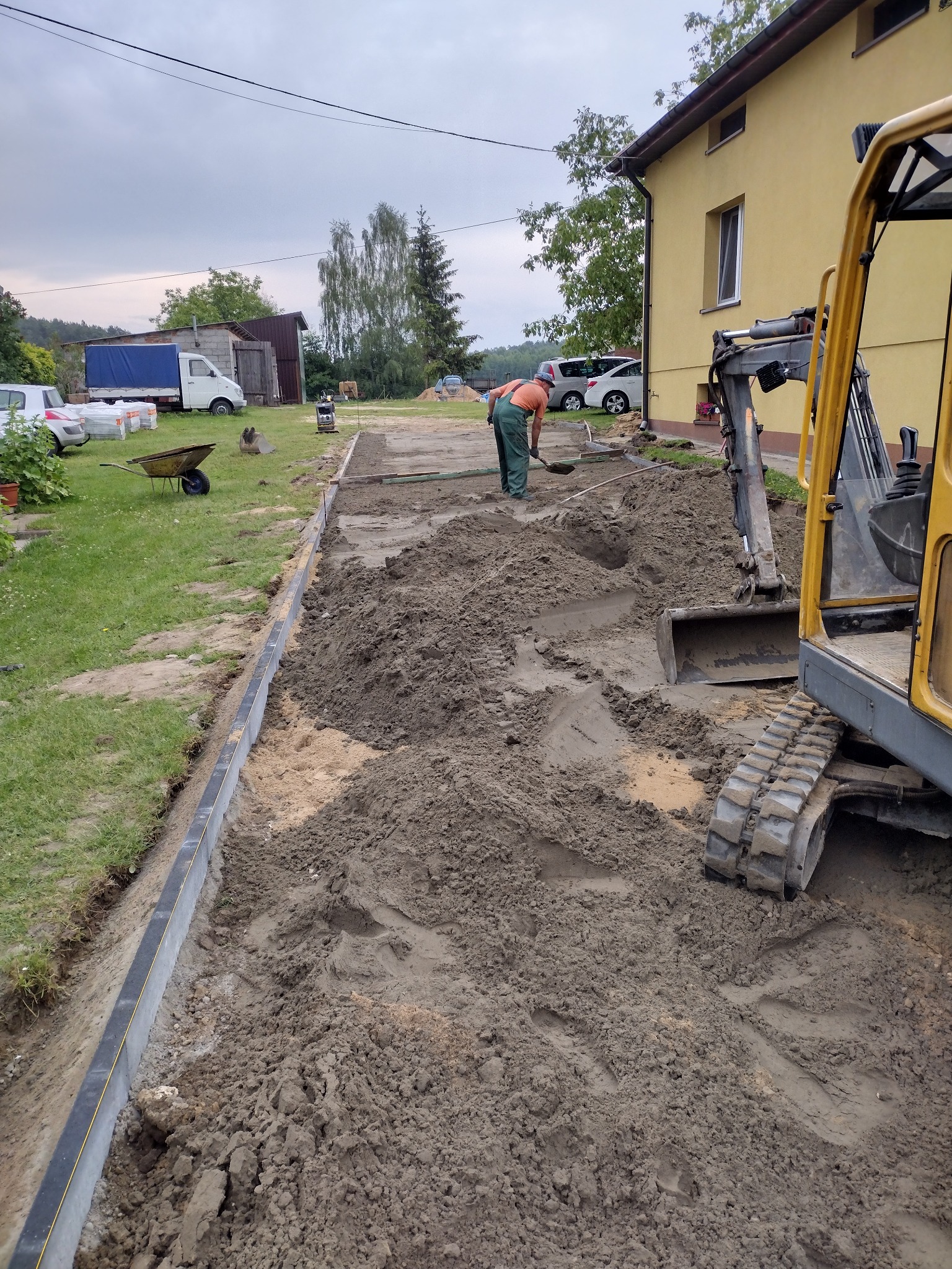 Pracownik wyrównuje ziemię łopatą wzdłuż krawężnika, w tle żółta ściana budynku i minikoparka, przygotowanie terenu pod budowę.