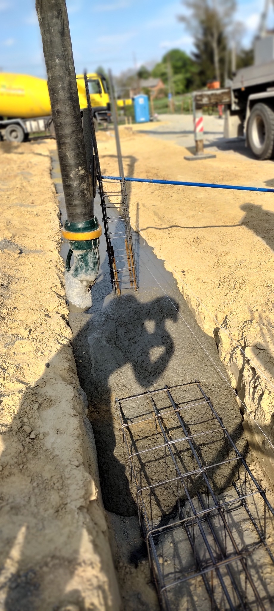 Wylewka betonu do wąskiego wykopu zbrojonego siatką stalową; w tle żółta betoniarka i cień operatora pompy do betonu.