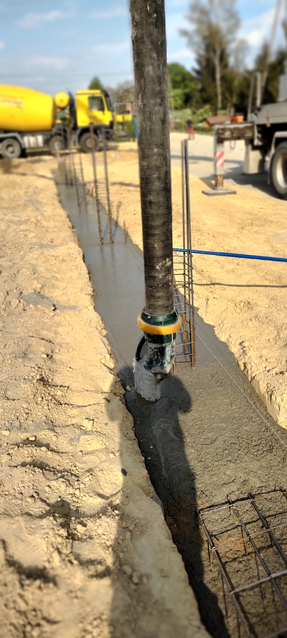 Wylewanie betonu do wąskiego wykopu zbrojonego stalą, widoczna pompa do betonu z żółto-zielonym łącznikiem i dwa żółte betonomieszarki w tle.