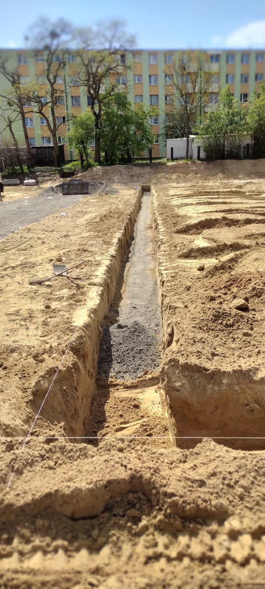 Wąski wykop w piaszczystej ziemi, częściowo wypełniony szarym żwirem, z budynkiem mieszkalnym w tle i narzędziami budowlanymi leżącymi obok.