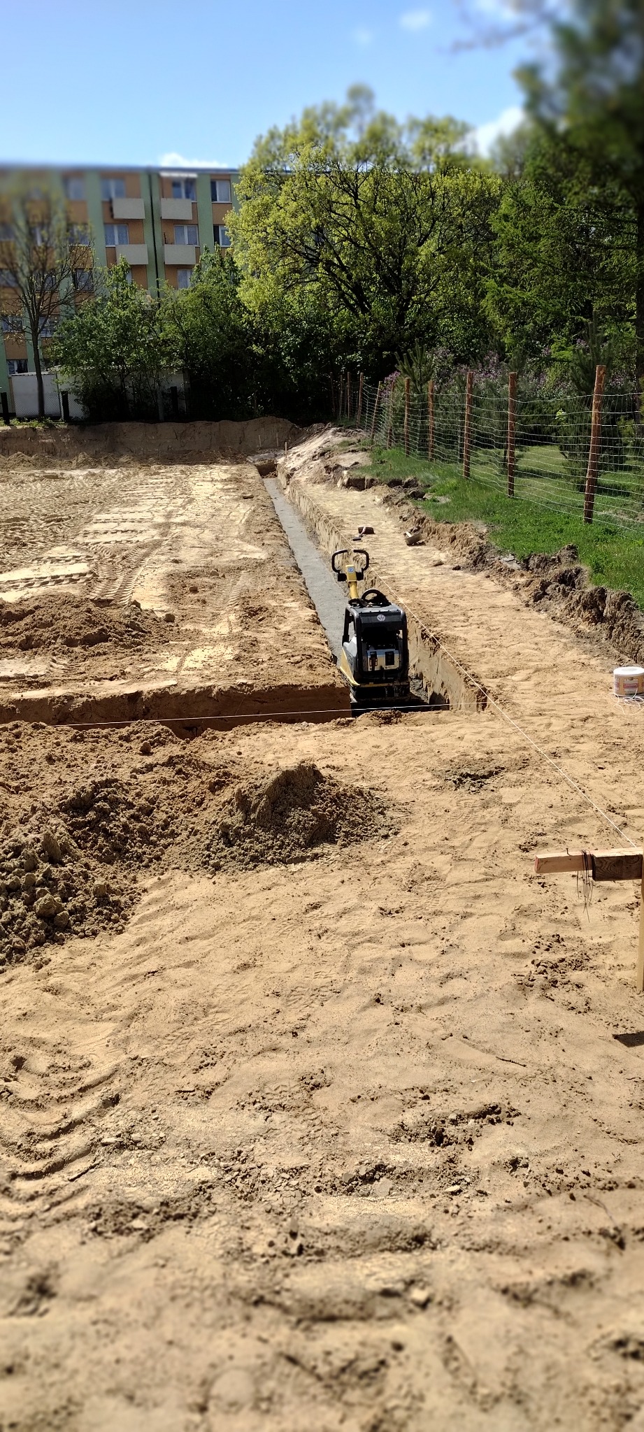 Wibro ubijak zagęszcza wąski wykop pod fundament na piaszczystym terenie, widoczne ślady opon i budynek w tle.