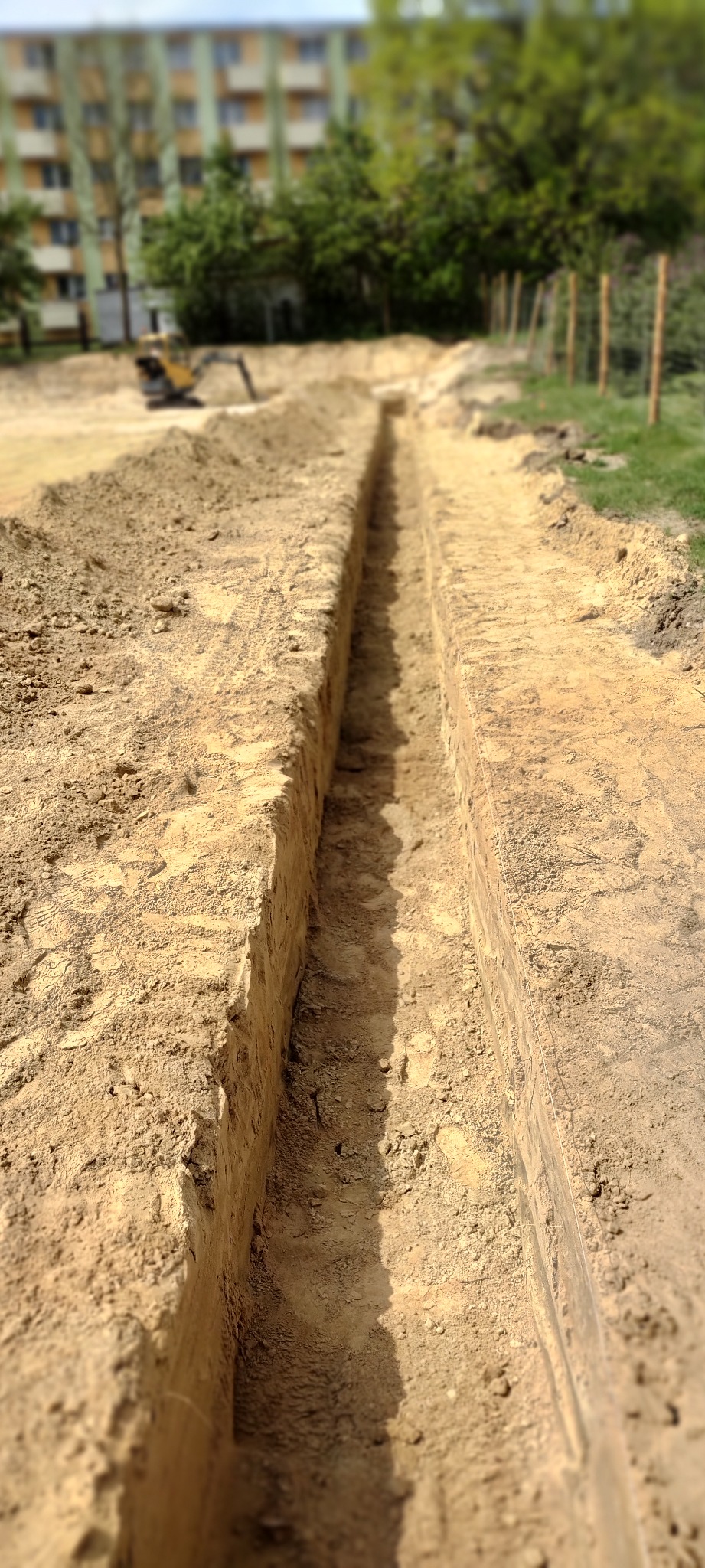 Długi, wąski wykop w piaszczystej ziemi, widoczny z perspektywy ziemi, z minikoparką w tle i budynkiem mieszkalnym w oddali.