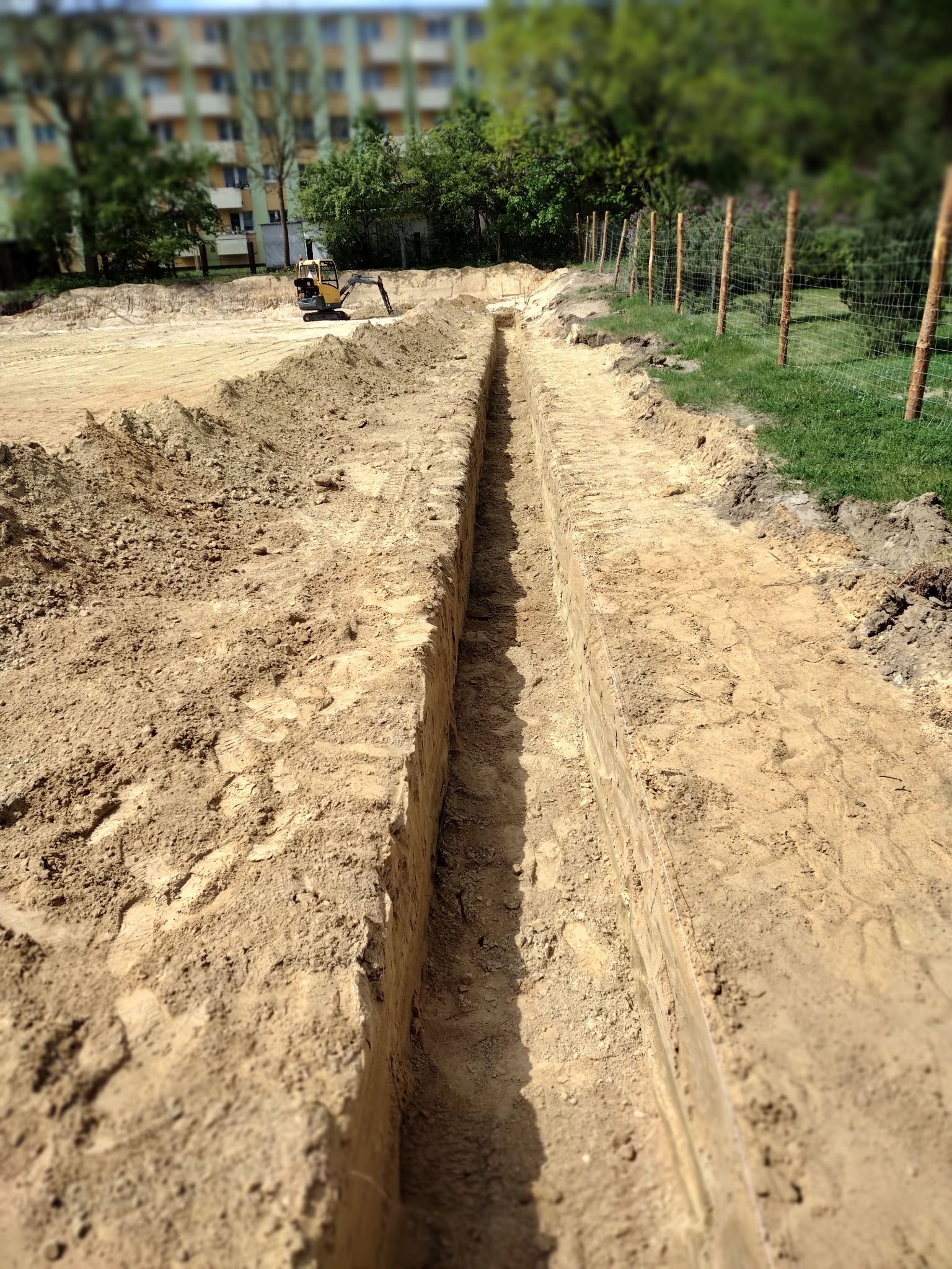 Wąski wykop liniowy w piaszczystej glebie, widoczny ślad po sznurze wyznaczającym linię, w tle mała koparka i budynek mieszkalny.