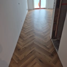 The Floors Łukasz Nykaza - Ułożona jodełka z paneli w jasnym korytarzu, widok w perspektywie, surowe ściany, w tle okno balkonowe z folią zabezpieczającą.