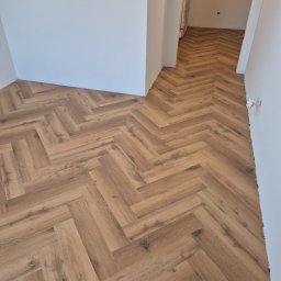 The Floors Łukasz Nykaza - Jasne panele ułożone w jodełkę francuską, widok z góry. Podłoga w odcieniach brązu, świeżo położona, przy białych ścianach i przejściu do kolejnego pomieszczenia.