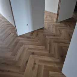 Ułożona podłoga z paneli w jodełkę, widok z góry, jasne panele drewnopodobne, surowy stan ścian, widoczne fragmenty instalacji elektrycznej.