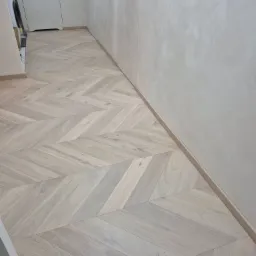 Jasna, drewniana podłoga ułożona we wzór jodełki francuskiej w korytarzu. Minimalistyczne wnętrze z białymi drzwiami i ścianami o fakturze betonu.