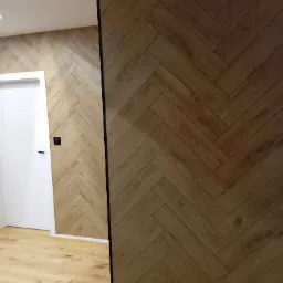 Wykończone wnętrze z panelami ściennymi ułożonymi w jodełkę, pasującymi do podłogi, białe drzwi po lewej stronie.