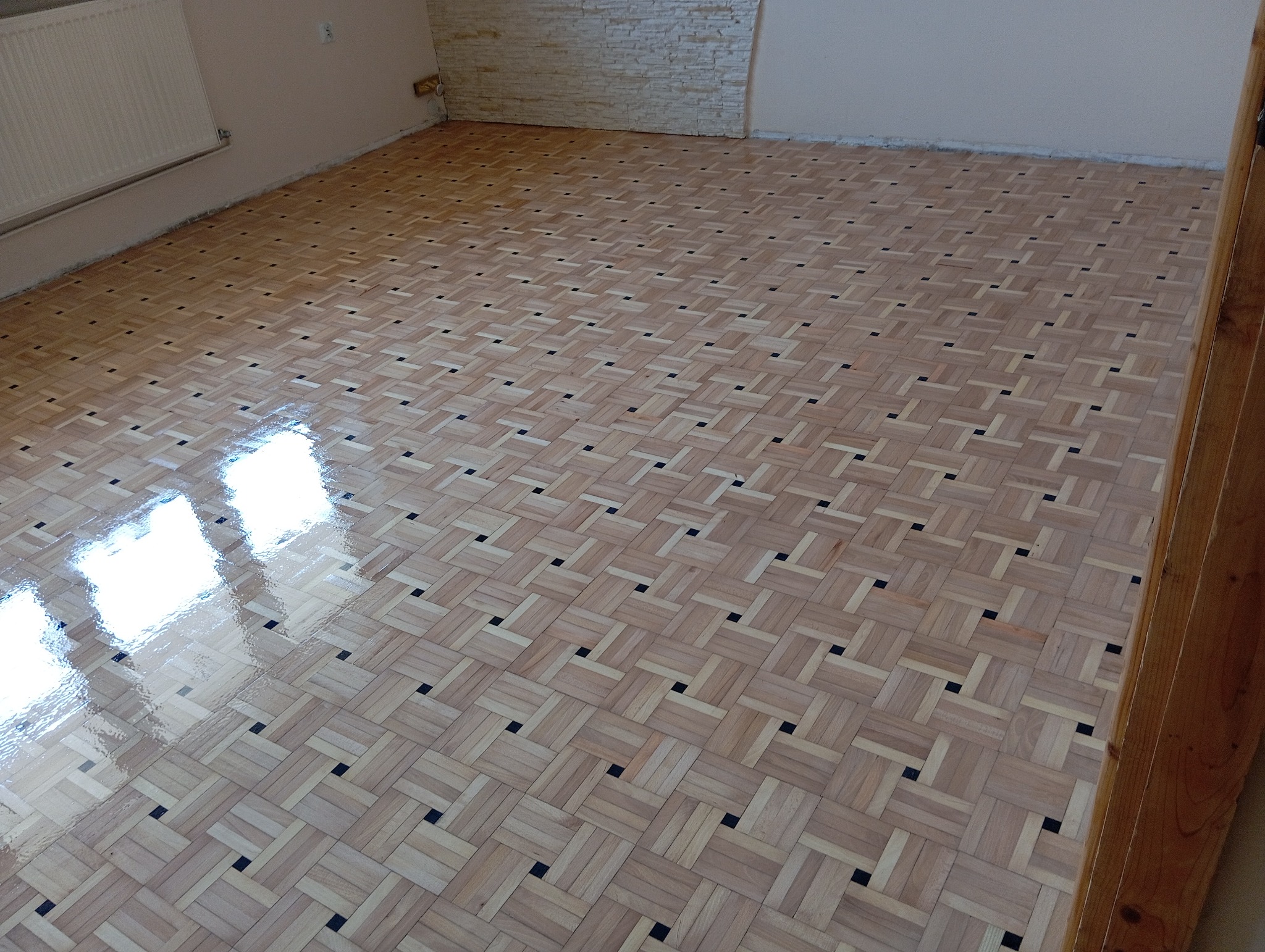 Świeżo położony, lakierowany parkiet w jodełkę z ciemnymi wstawkami, odbijający światło w pustym pokoju z grzejnikiem i fragmentem dekoracyjnej ściany.